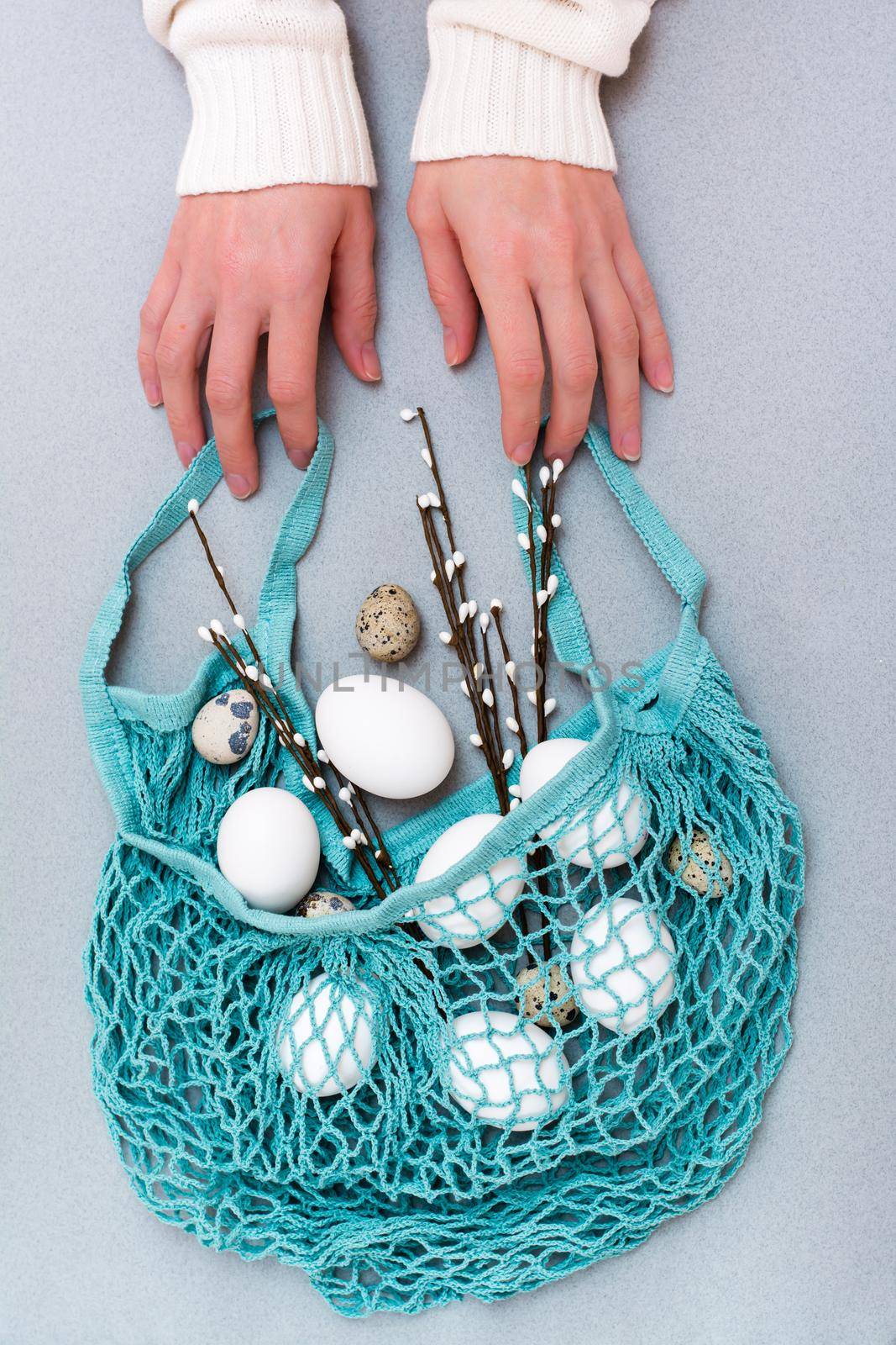 Happy Easter. Female hands hold a blue string bag with chicken and quail eggs and pussy willow branches on a gray background by Aleruana