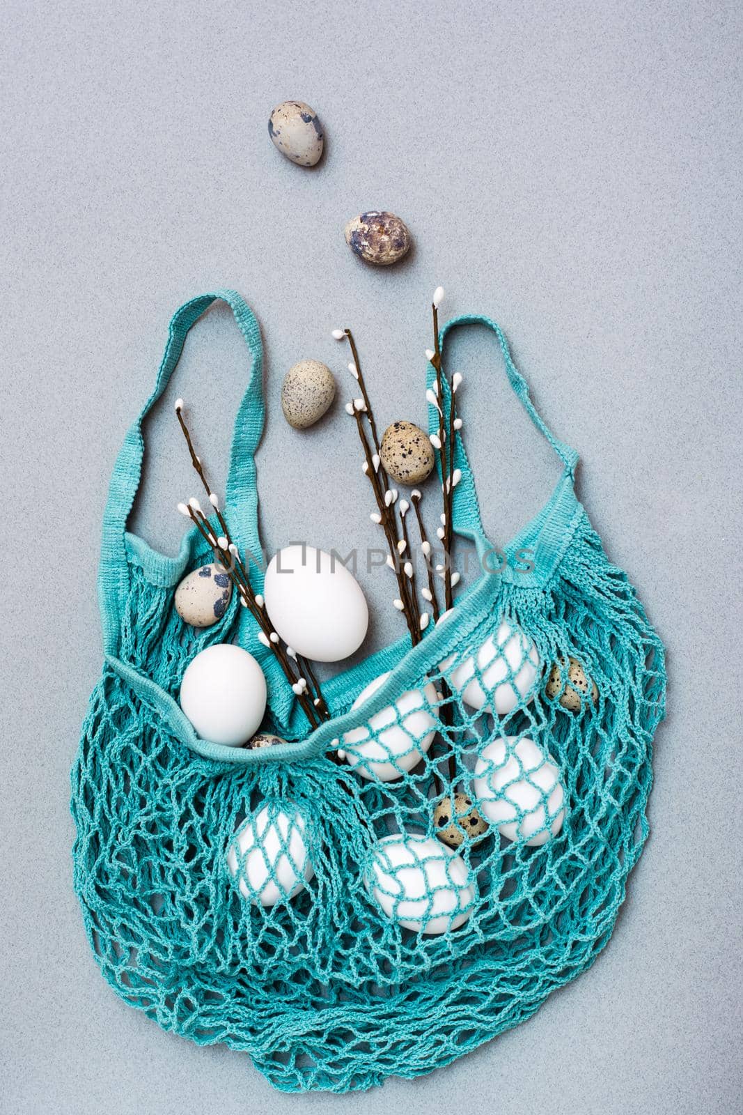 Happy Easter. Chicken and quail eggs falling into a blue mesh bag with pussy willow branches on a gray background by Aleruana