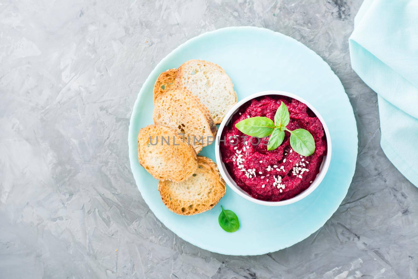 Homemade baked beetroot hummus in a bowl with sesame seeds and basil and baked small toast on a plate on the table. Top view