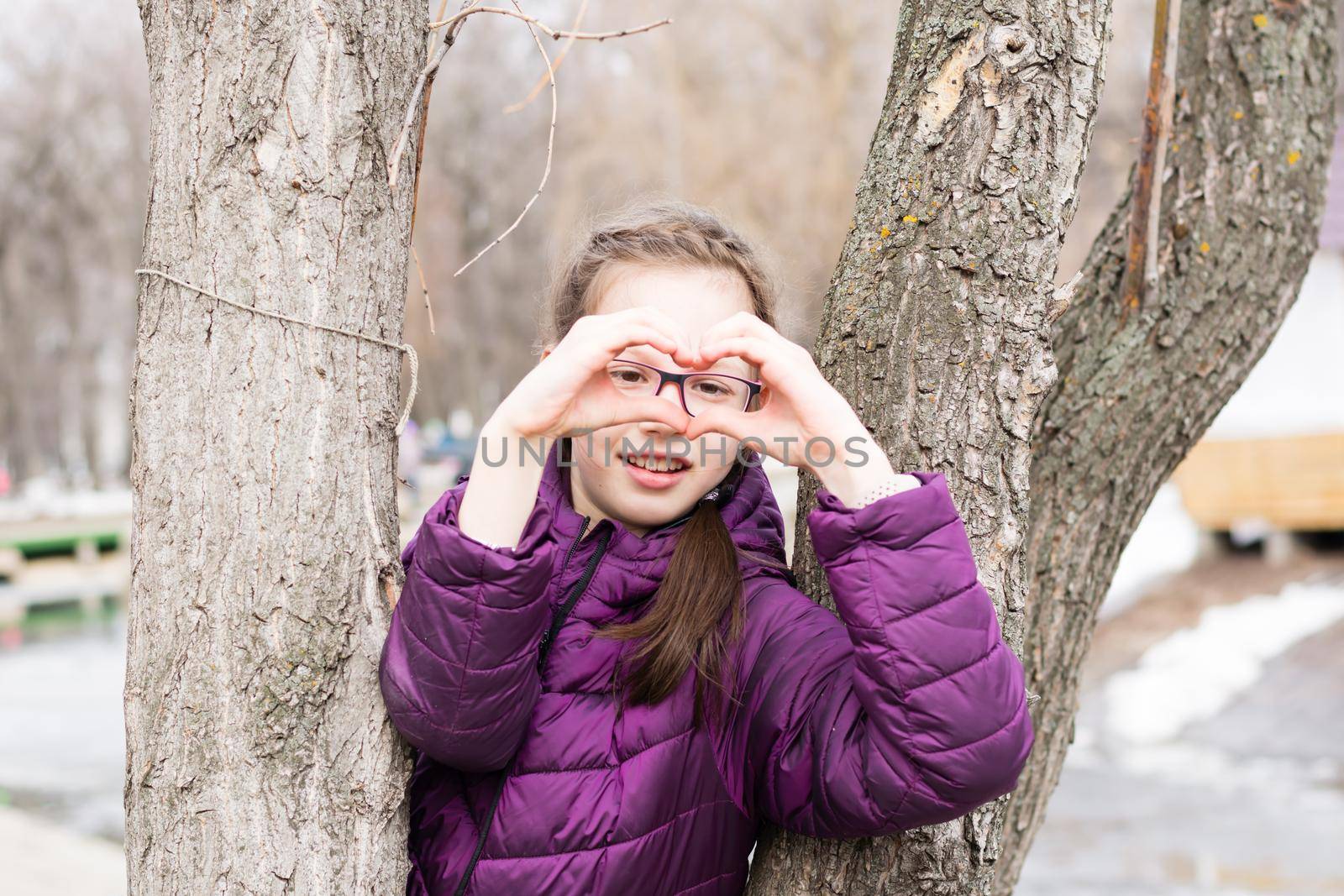 Young attractive girl in glasses makes a heart shape with her palms in front of her face in a city park in early spring by Aleruana
