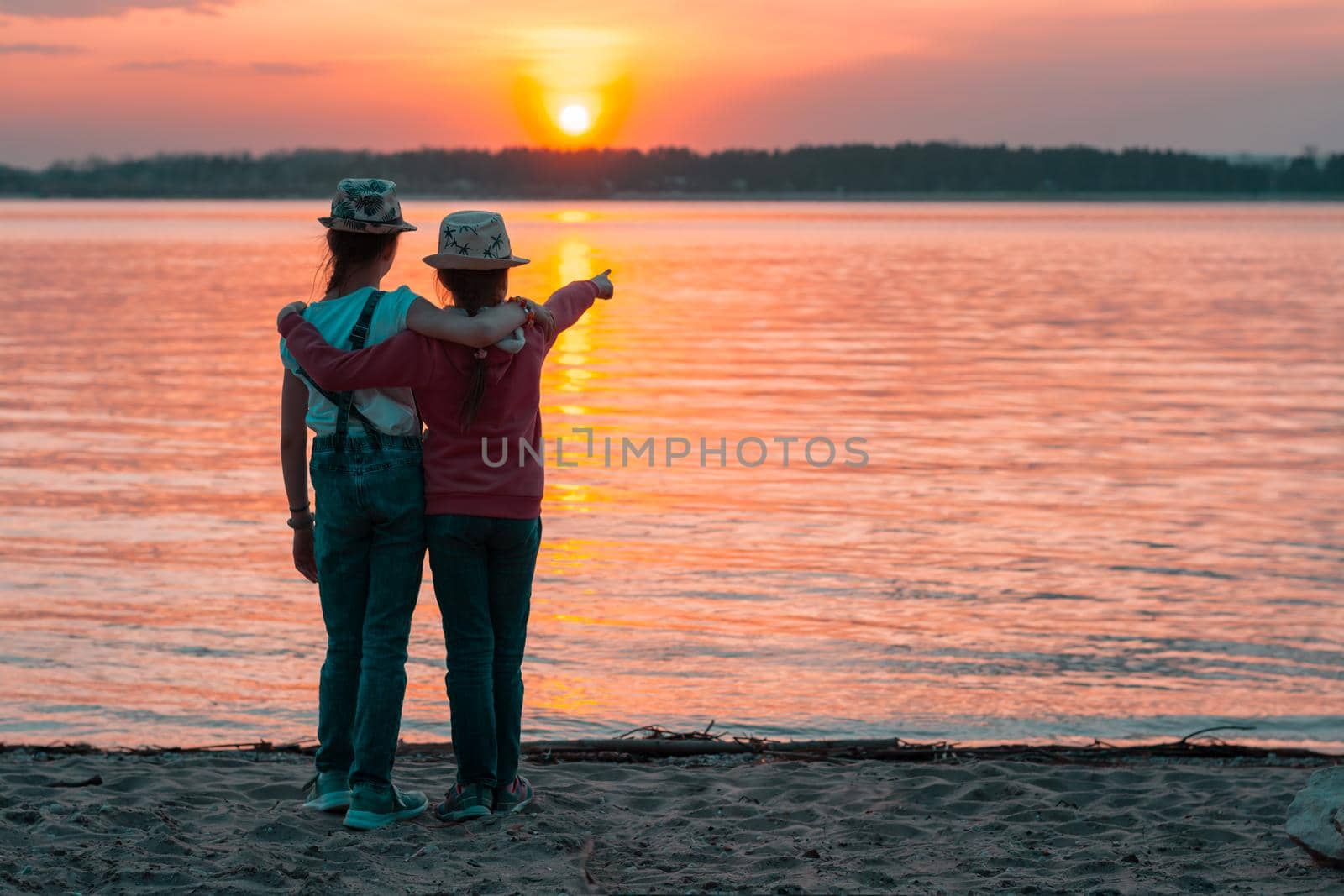 Two girls sisters stand on the sand on the bank of the river in the sunset rays of the sun and look at the opposite bank. Local tourism by Aleruana