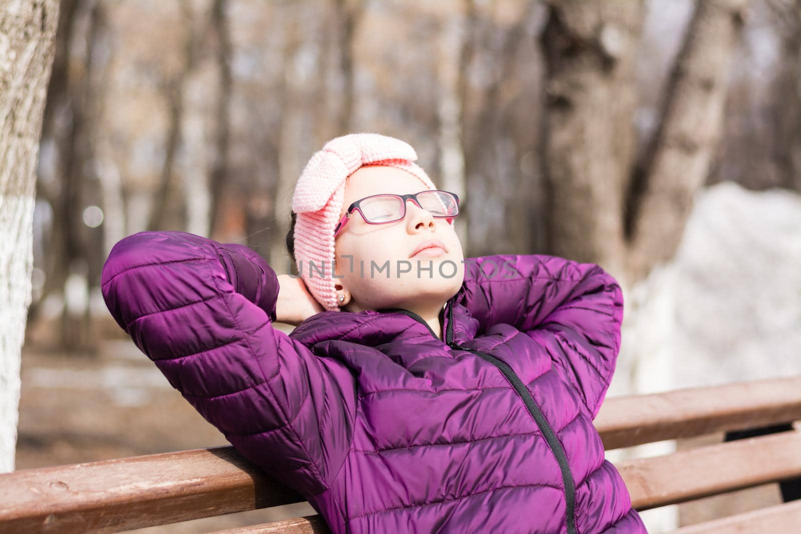 A girl in a jacket and glasses on a bench in a city park in early spring put her face to the rays of the sun and enjoys the first warmth