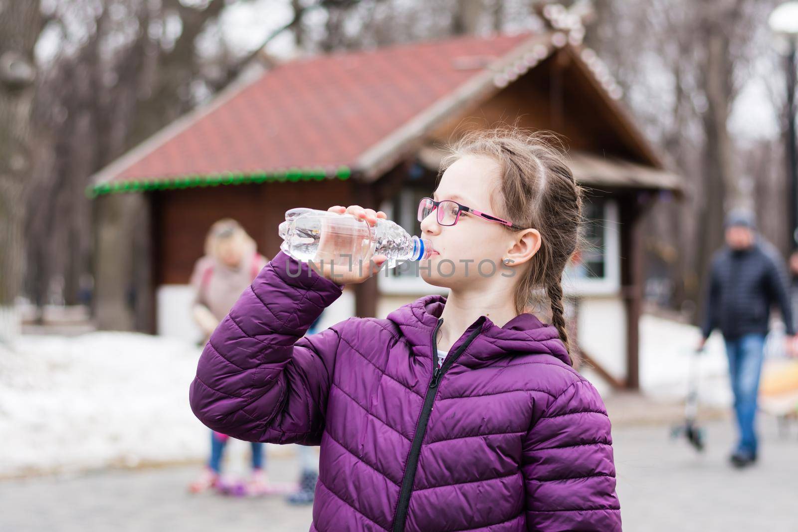 A cute girl in glasses drinks water from a bottle bought in a food truck in a city park. Takeaway food