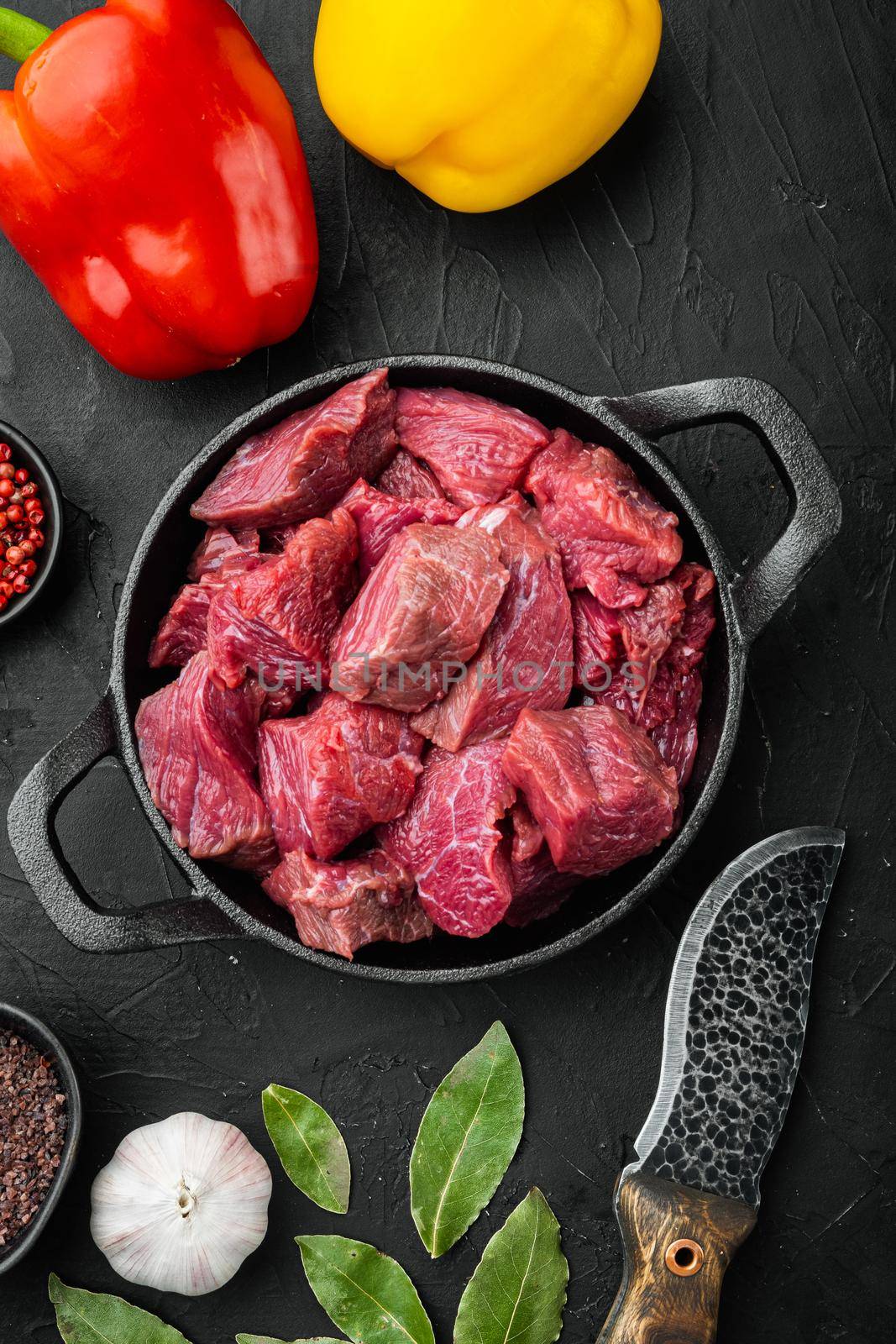 Fresh beef chunks served on table with ingredients ready for cooking with sweet bell pepper, in cast iron frying pan, on black stone background, top view flat lay by Ilianesolenyi