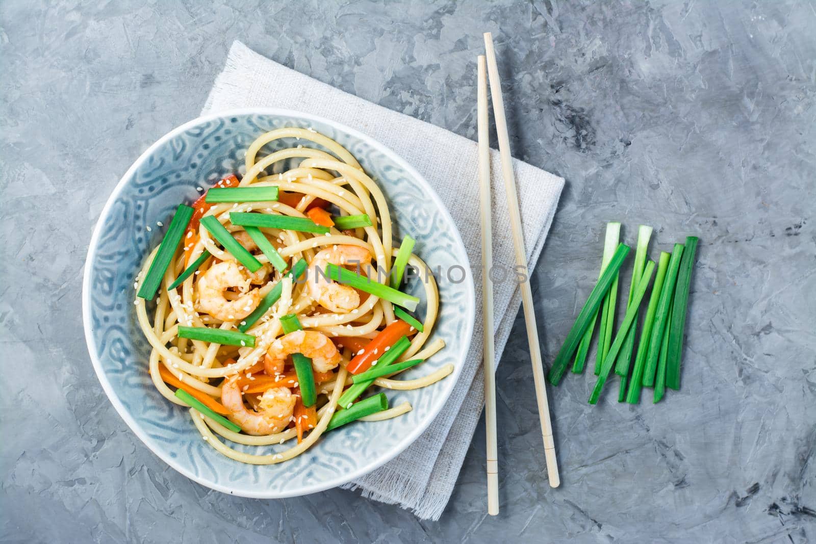 Udon noodles and wok with shrimps, peppers and onions in a plate on a napkin and chopsticks on the table. Chinese food. Top view by Aleruana