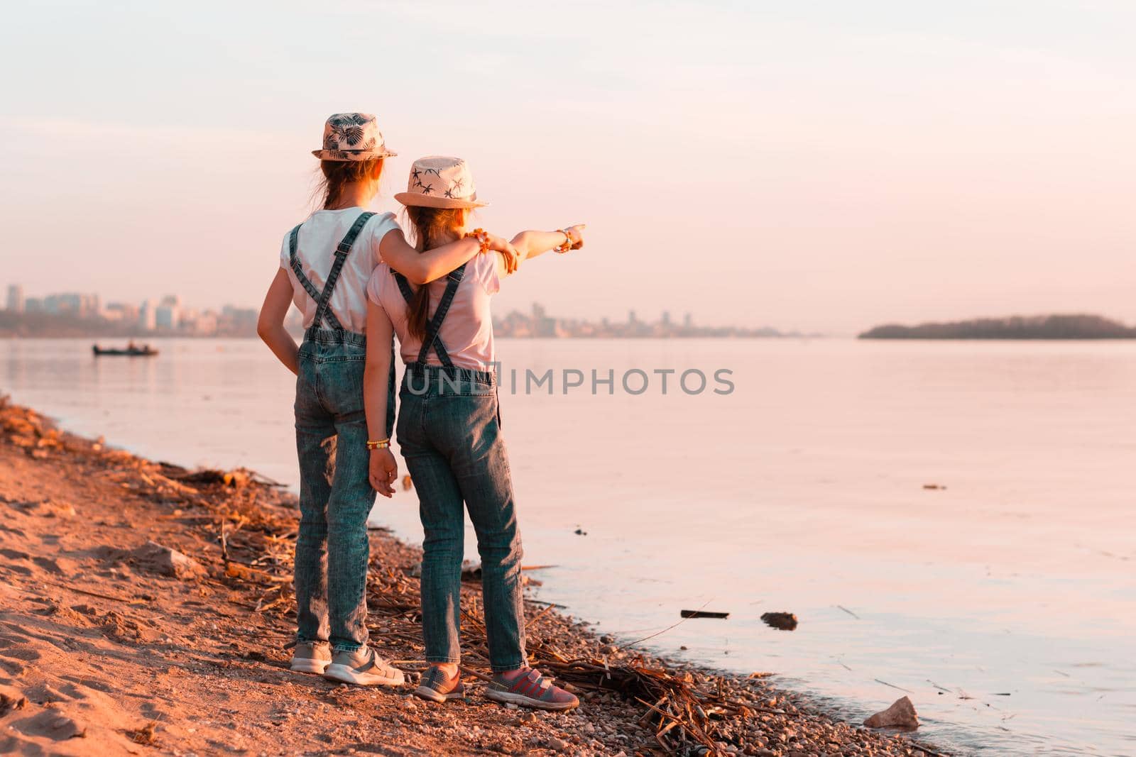 Two girls sisters stand on the banks of the river at sunset and look into the distance. Local tourism by Aleruana