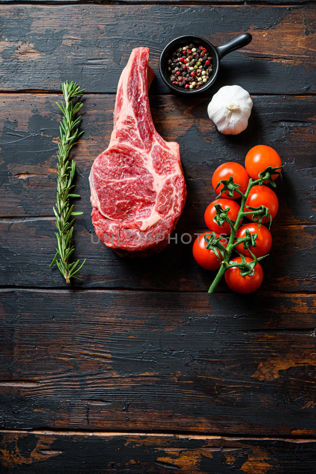 Raw uncooked black angus beef Ribeye Steak, Bone-Incloseup with tomatoes garlic herbs and peppercorns rosemary on wooden dark old rustic table planks with space for text layflat