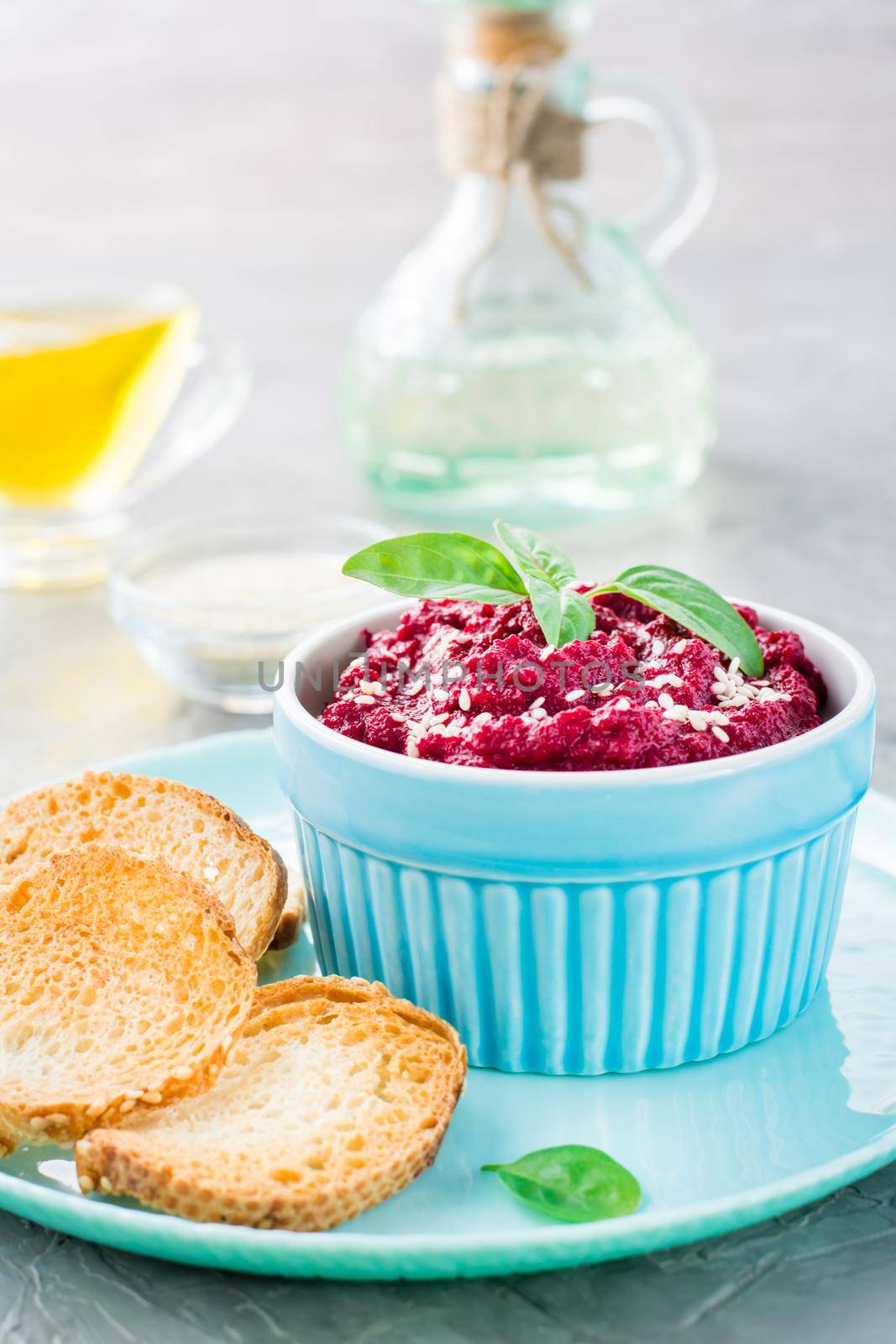 Homemade baked beetroot hummus in a bowl with sesame seeds and basil and baked small toast on a plate on the table. Vertical view