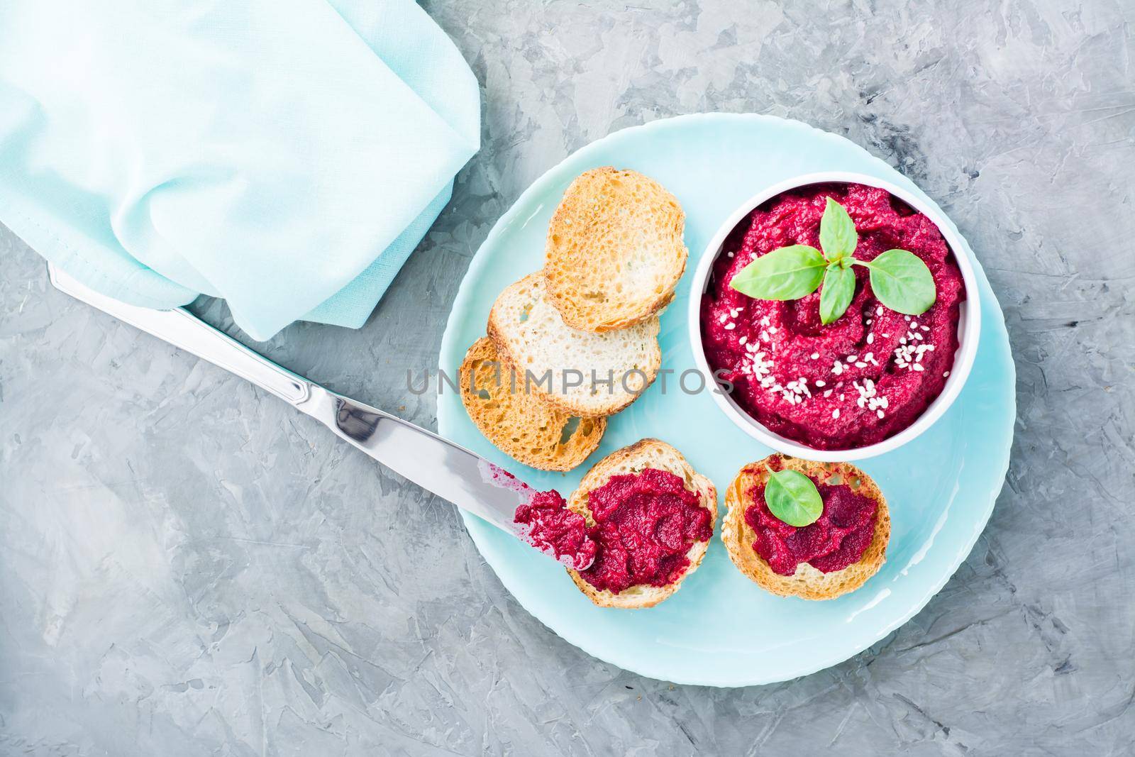 Homemade bruschetta with beetroot hummus on small toast on a plate and a table knife on the table. Top view