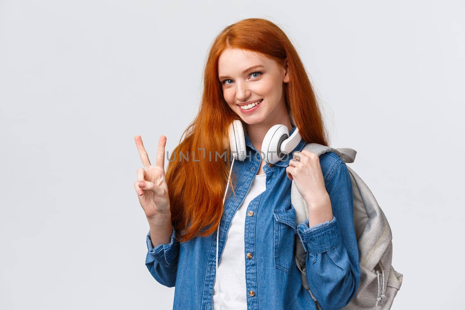 Lovely flirty and cheeky redhead college girl, wear headphones over neck and backpack, showing peace sign and smiling alluring camera, standing happy over white background enjoy student life.