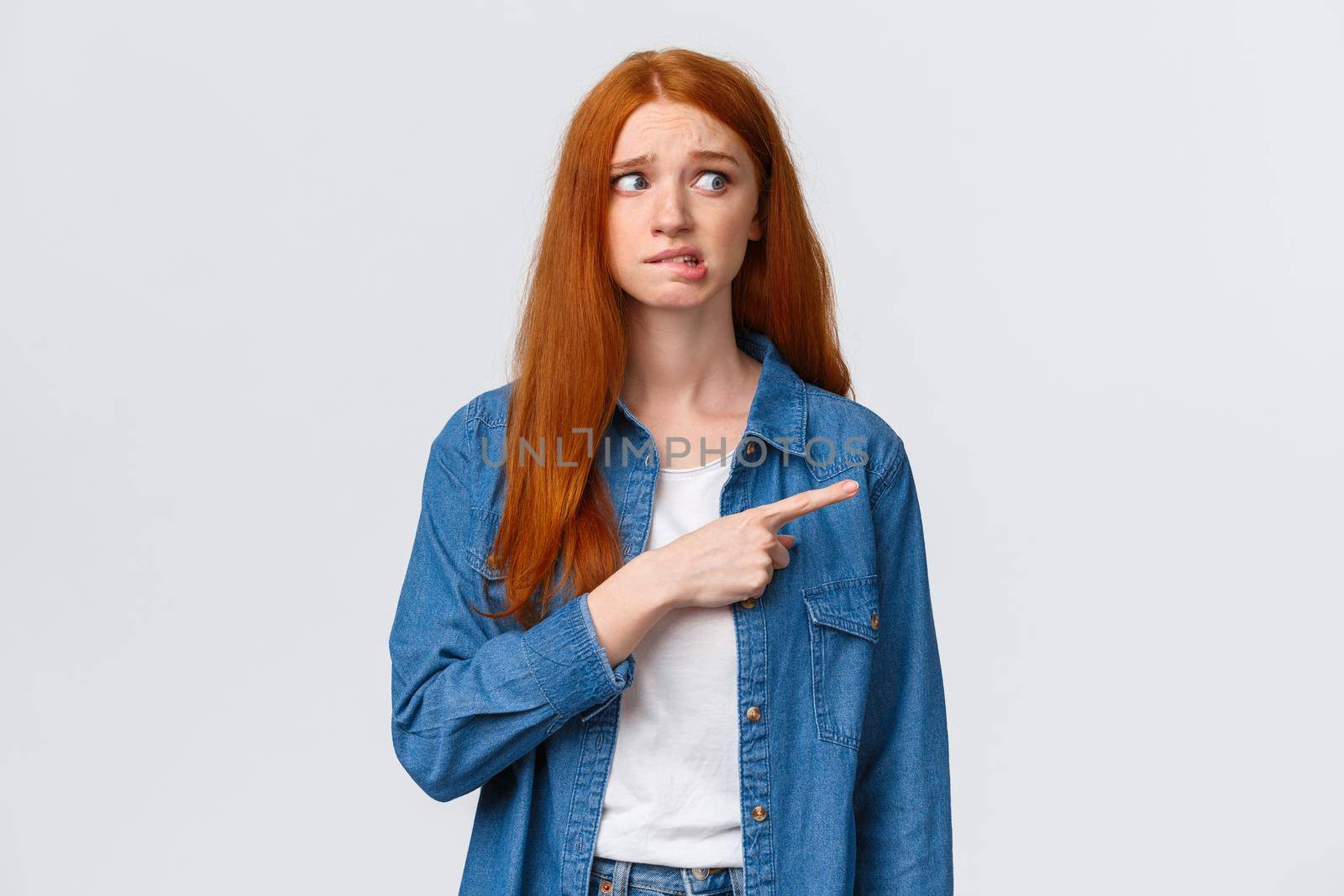 Awkward and nervous cute timid redhead teenage girl having fear or embarrassing thing say, pointing looking right anxious, biting lip and frowning, confessing in crime, feel guilty.