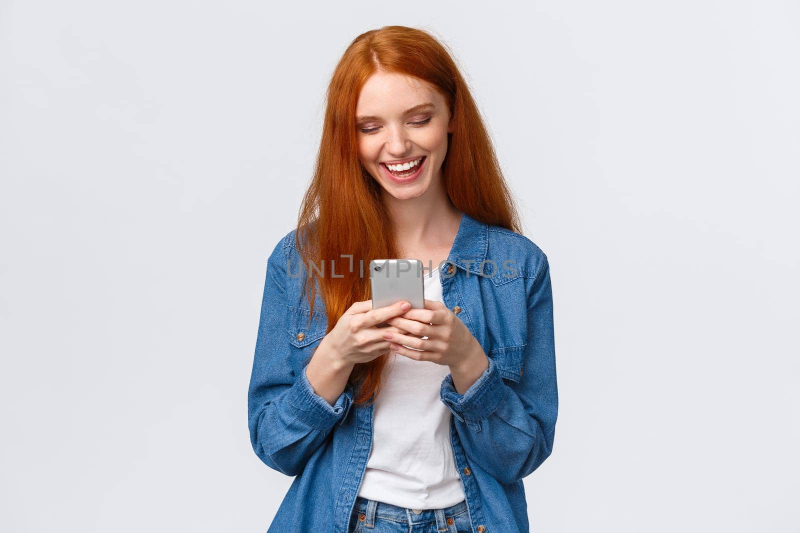 Chatting, communication and mobile concept. Cheerful redhead smiling girl texting friend, laughing as watching funny vlog, looking smartphone display happy, standing white background.