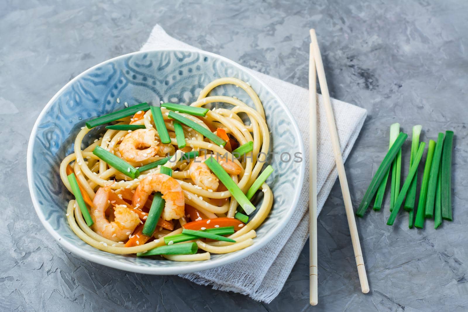 Udon noodles and wok with shrimps, peppers and onions in a plate on a napkin and chopsticks on the table. Chinese food