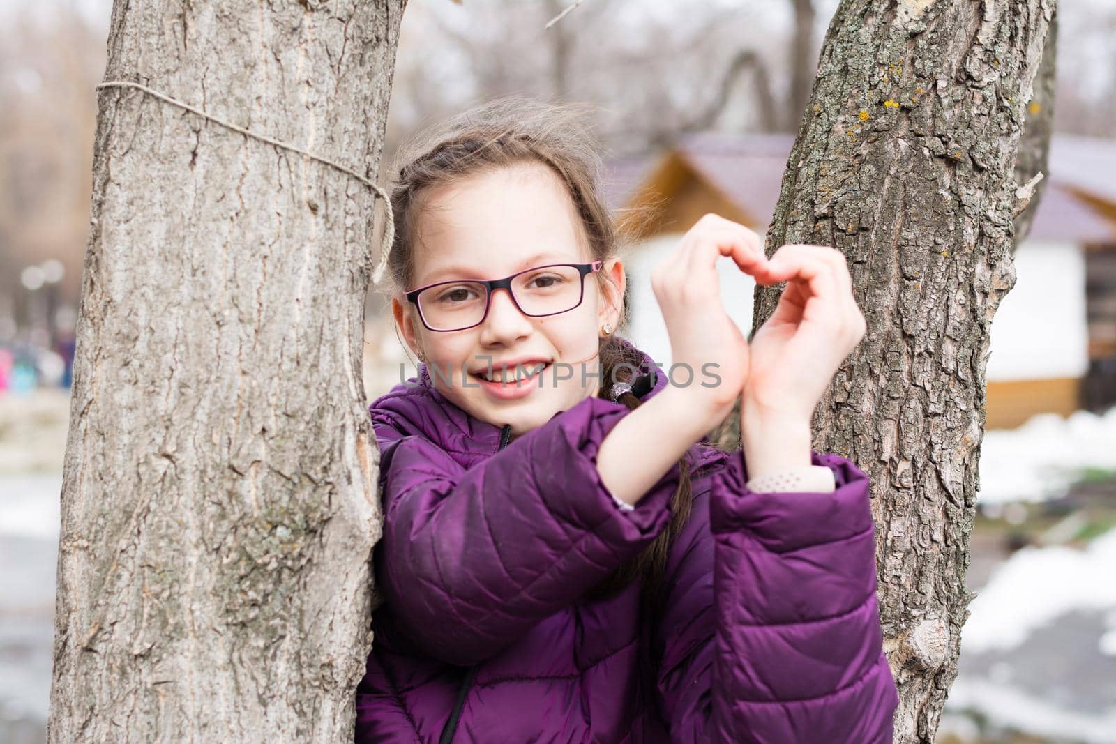 Young attractive girl in glasses makes a heart shape with palms in a city park in early spring by Aleruana