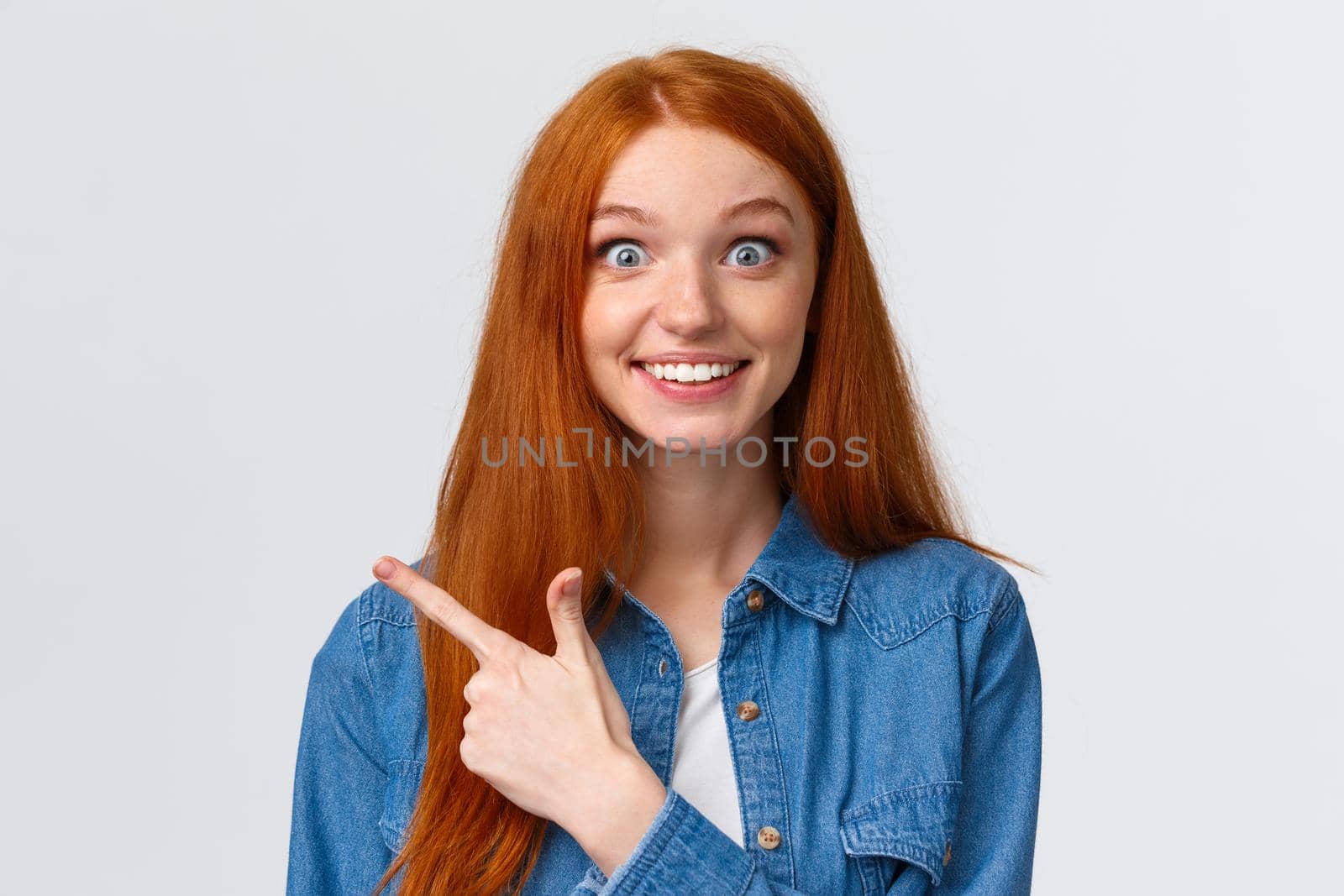 Excited and impressed, curious thrilled smiling redhead girl with long foxy, ginger hair and blue eyes, stare amused camera with wondered grin, pointing finger left, found out awesome news.