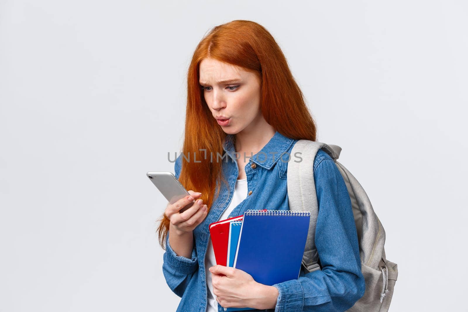 What is that. Confused and displesaed. intense troubled redhead teenage, college girl with backpack and notebooks walking university and reading strange text on smartphone, white background.
