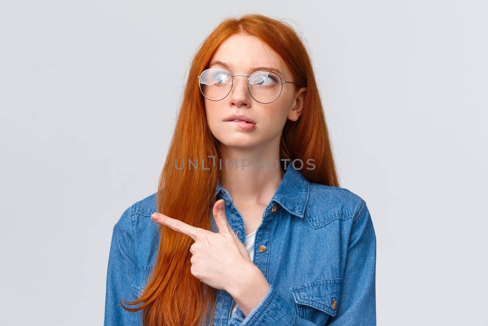 Unsure smart and cute redhead teenager in glasses, female student biting lip curious looking and pointing left, thinking, deciding what pick, making choice, standing over white background.