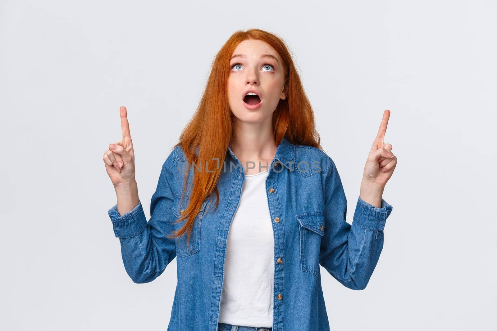 Waist-up portrait amazed and impressed, curious redhead girl checking out awesome game in store, drop jaw, looking and pointing up speechless, gasping amused, stand white background.