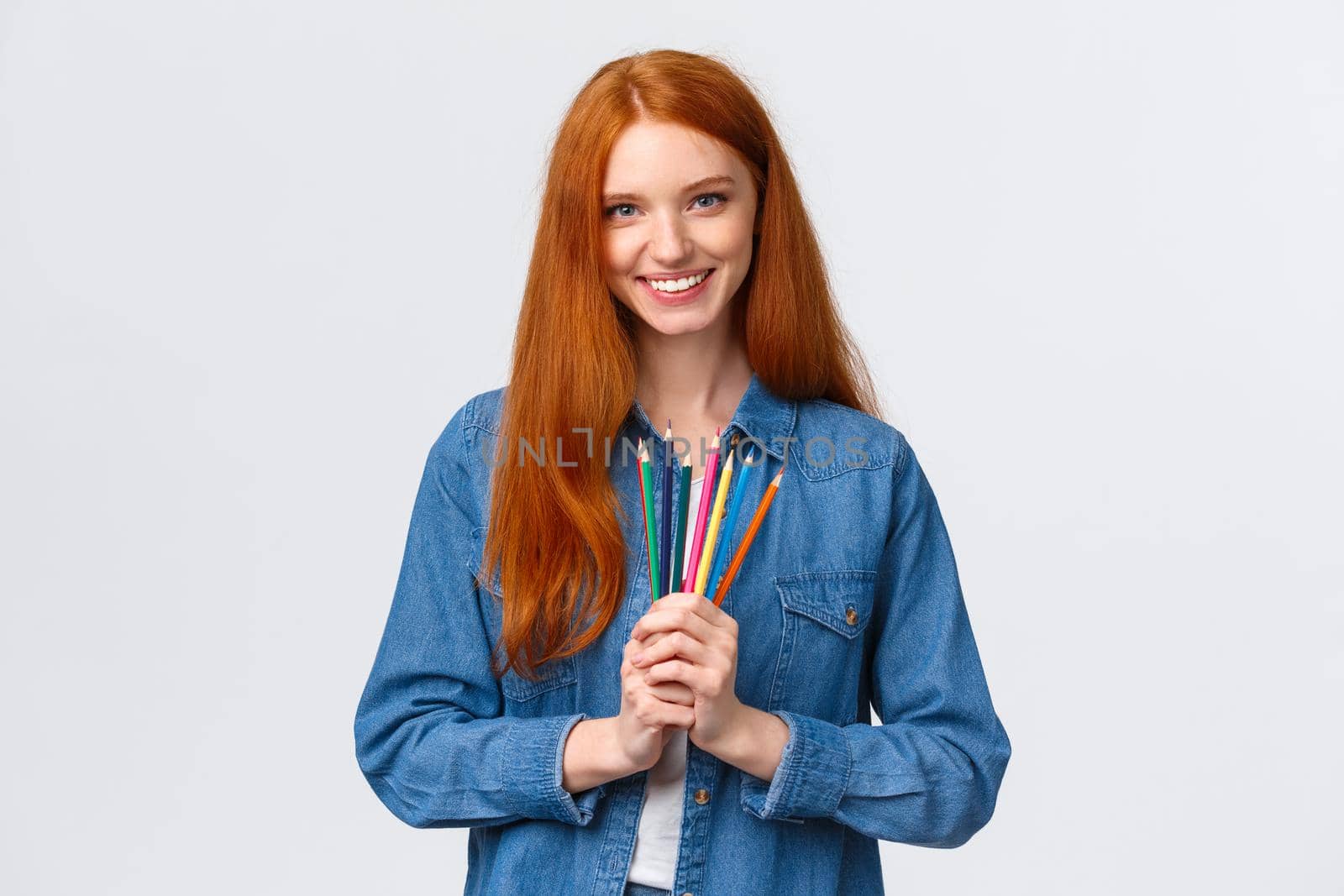 Time to brighten life. Attractive confident charismatic redhead girl in denim shirt, looking camera with pleased smile, holding colored pencils, standing white background drawing, attend courses.