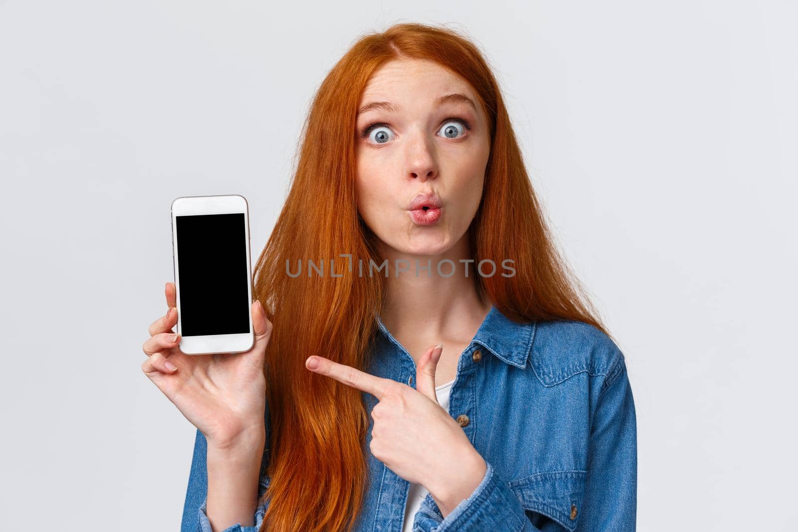 Curious and amused lovely redhead woman with long red hair, folding lips intrigued and excited, discussing new app, photos of classmate with new car, pointing finger smartphone, gossiping.