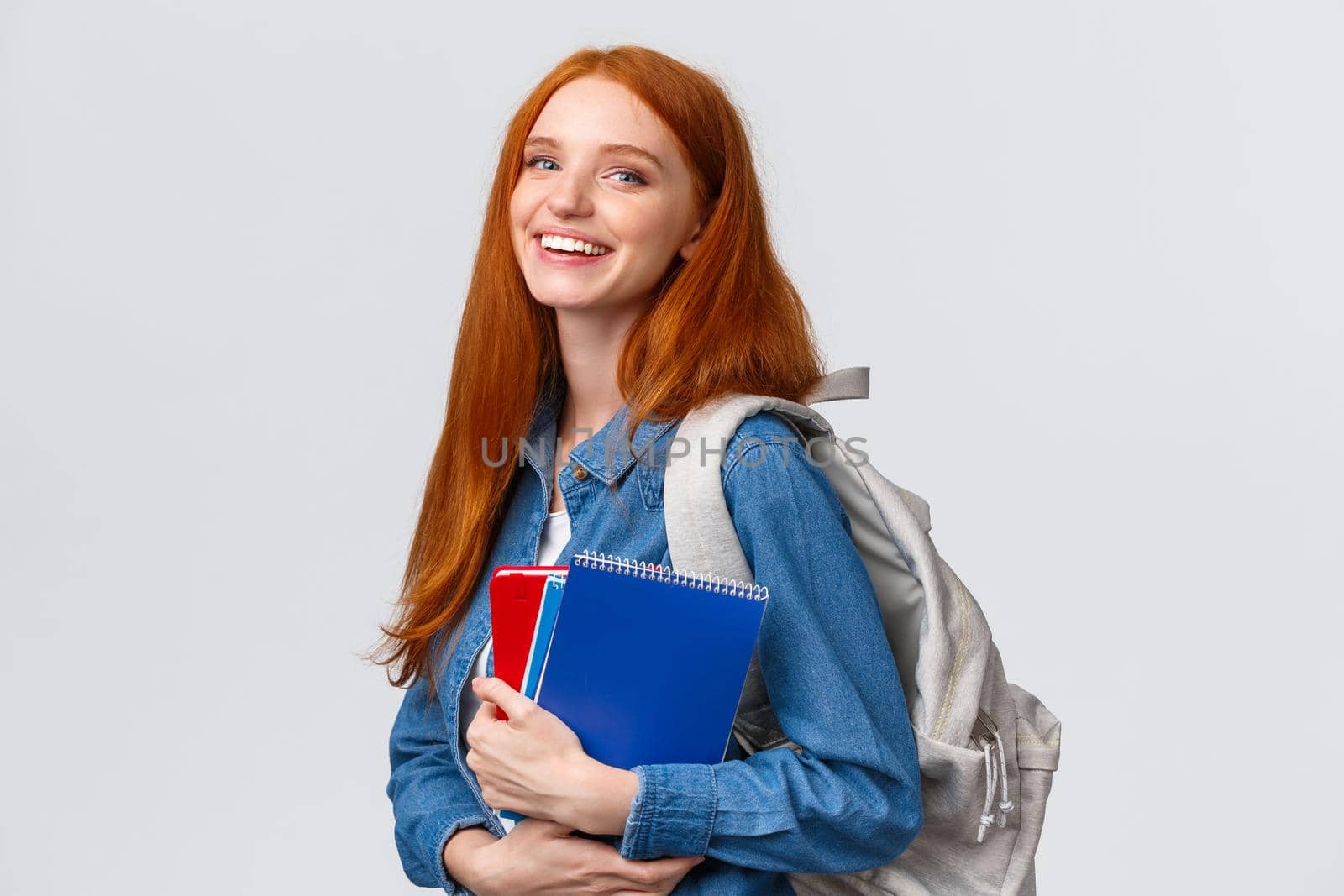 Teenagers, students and education concept. Cheerful lovely redhead female studying, going to univeristy or college, smiling carefree holding backpack and notebooks, standing white background.