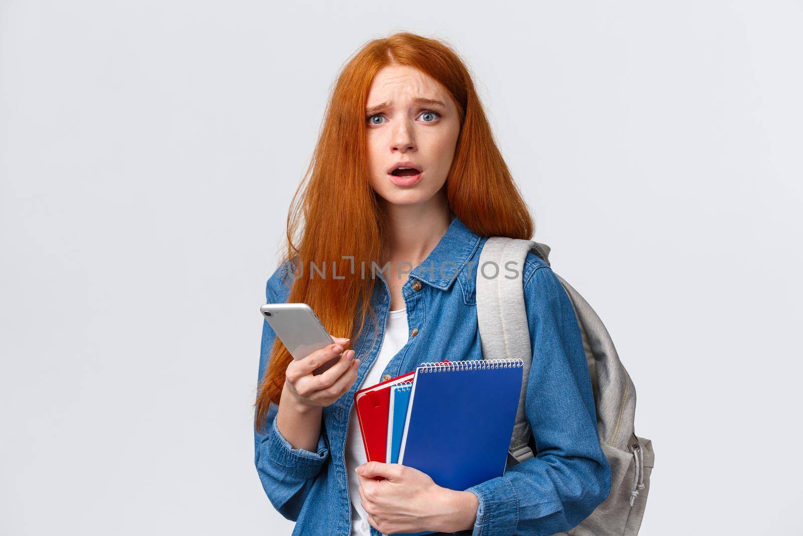 Uneasy and sad gloomy cute redhead female looking troubled camera, reading something worrying on smartphone display, holding mobile, backpack and notebooks as in university.