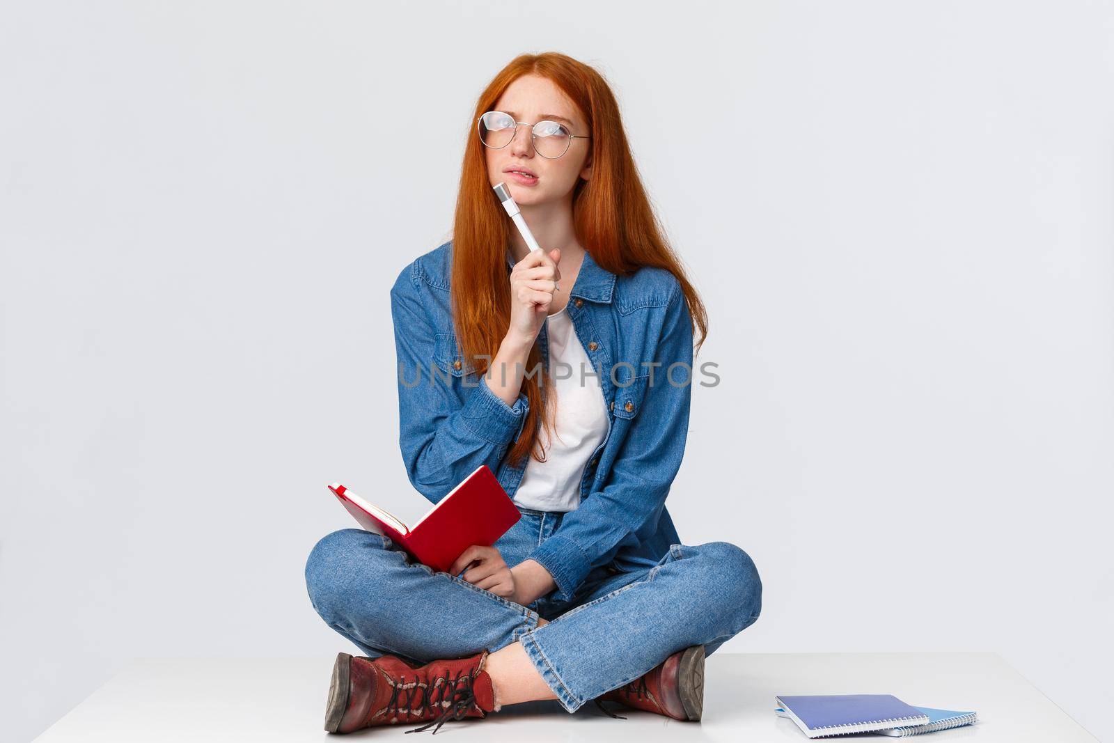 Concentrated, thoughtful and creative young female genius with red hair, in glasses, looking perplexed and thinking over solution to task, writing homework sitting on floor with notebook and pen.