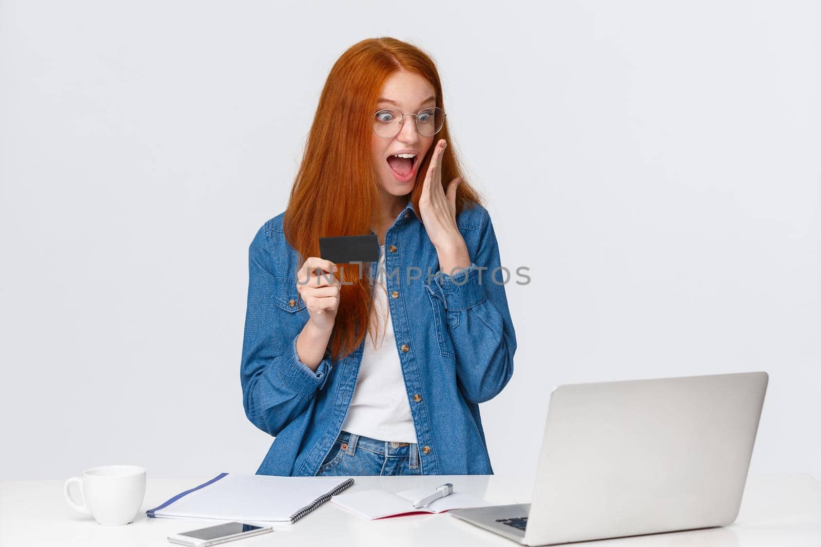 Excited and amused, happy redhead woman seeing something awesome on screen laptop, drop jaw gasping astonished, touch face, holding credit card, standing white background impressed.