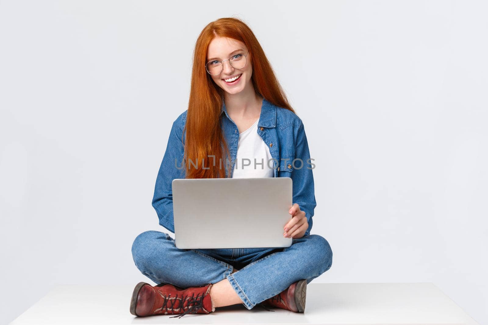 Cheerful sassy redhead female student, college girl in glasses, sitting on crossed legs with laptop, working freelance, prepare project awat from office, remote over white background.