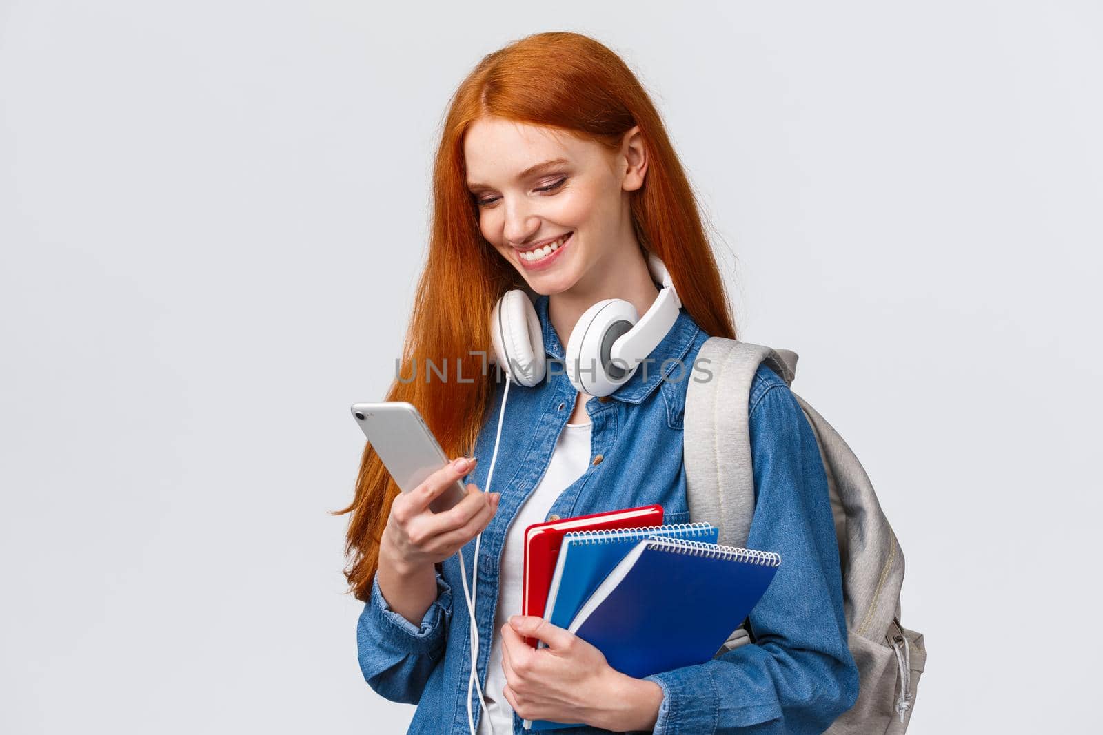 Teenagers, college and education concept. Attractive redhead female, univeristy student with headphones, backpack and notebooks, answer girlfriend message, queer love in air.