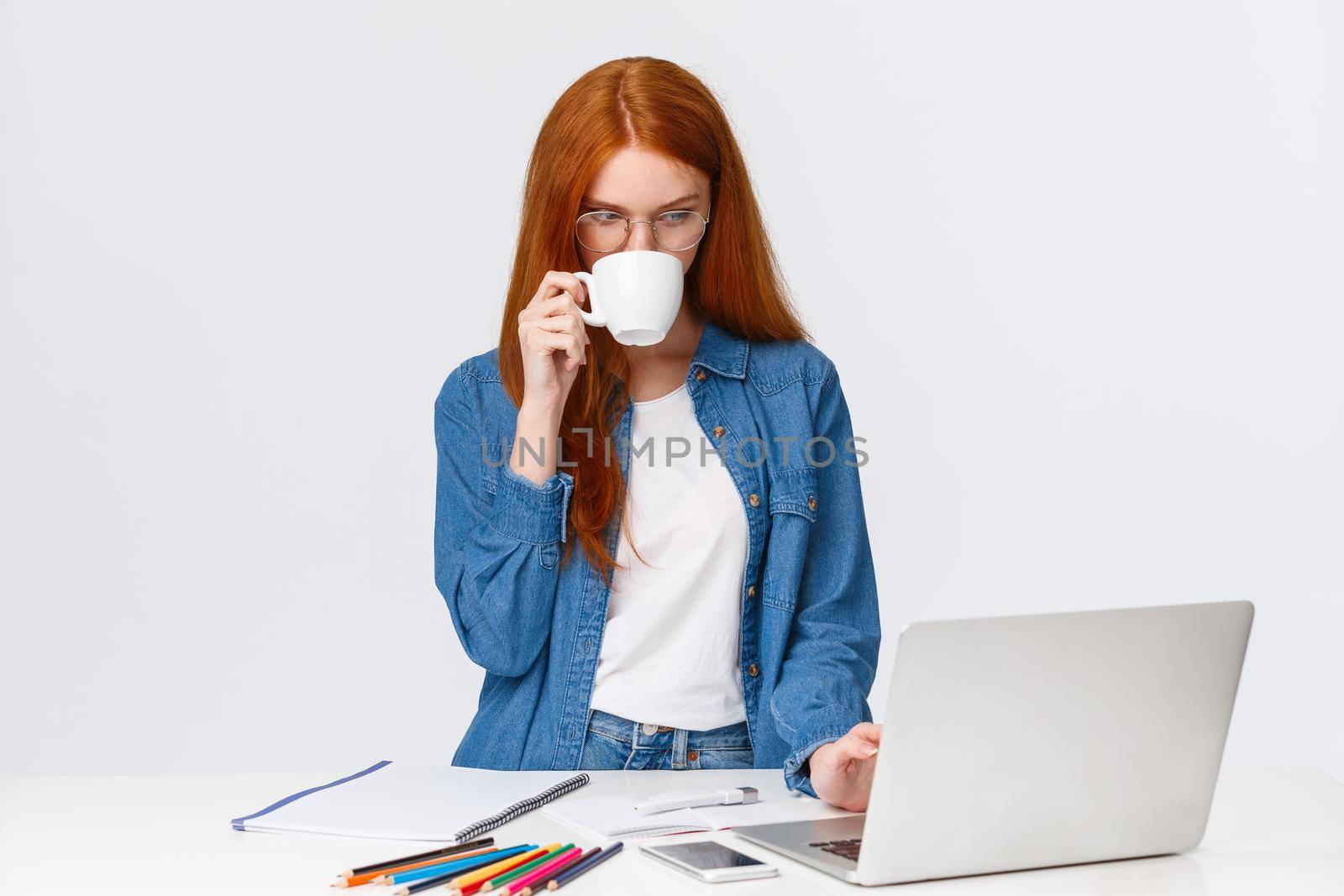 Serious-looking determined redhead female designer, office worker have no time for coffee break, sipping from cup, looking at laptop as working over important project, stand white background.