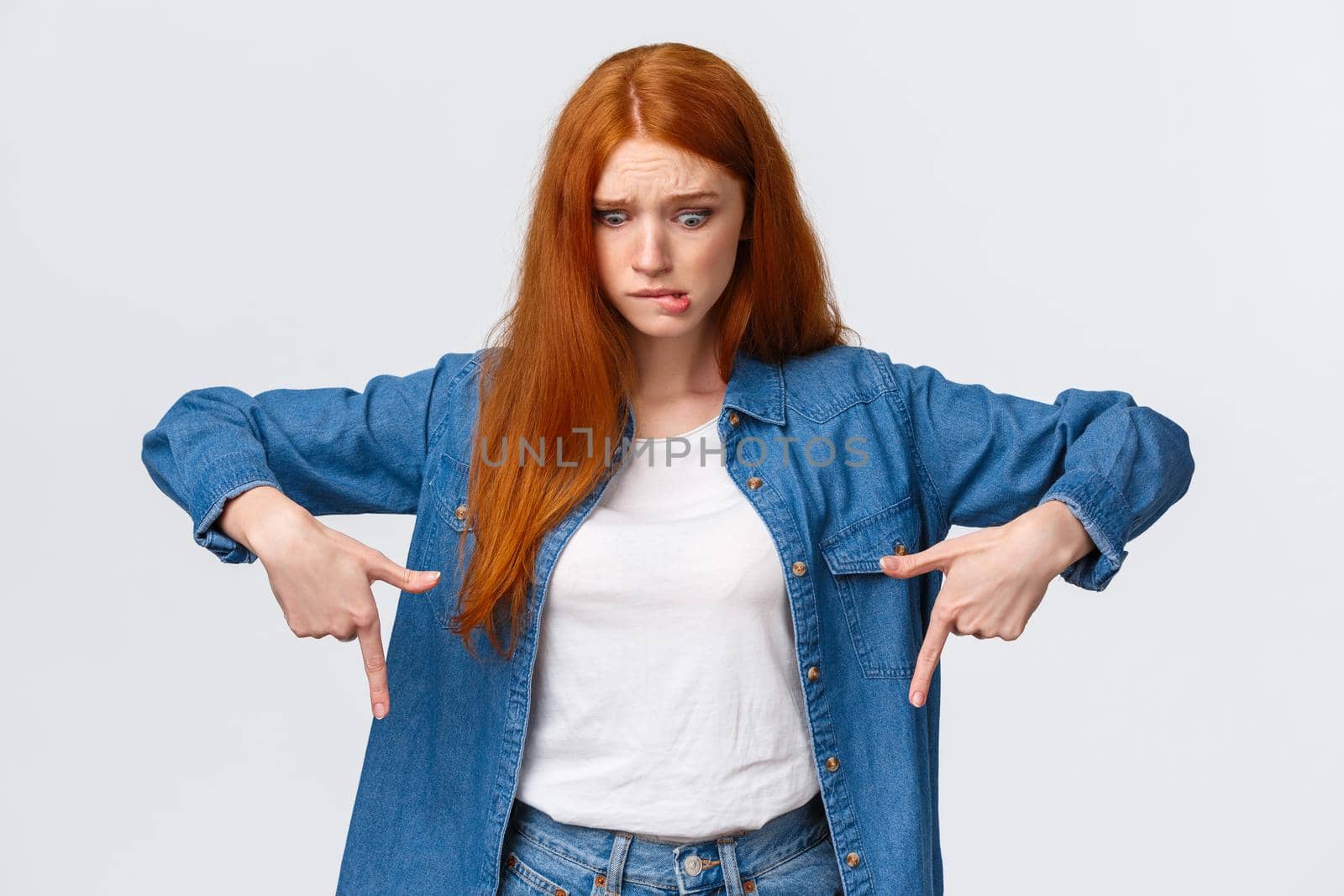 Embarrassed and worried, anxious cute redhead girl dropped something, feeling nervous, biting lip and staring gloomy down, pointing bottom advertisement, standing white background.