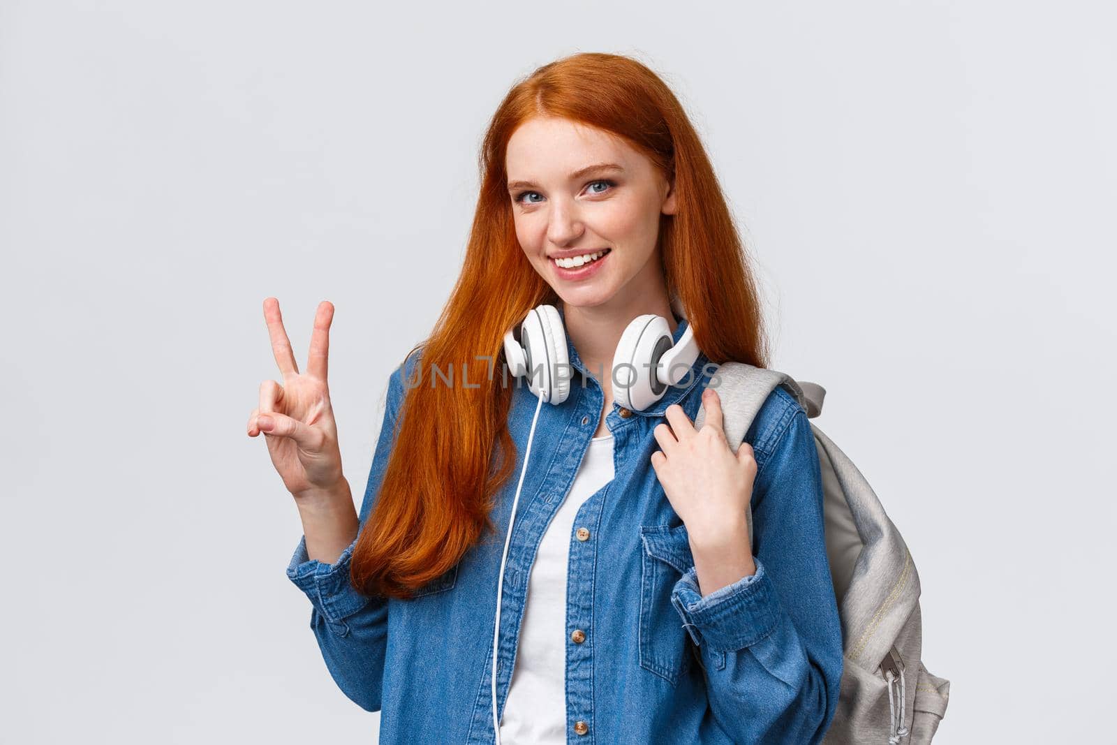 Lovely charismatic caucasian redhead girl with backpack, headphones over neck, showing peace sign and smiling delighted and joyful camera, standing white background, heading part-time job.