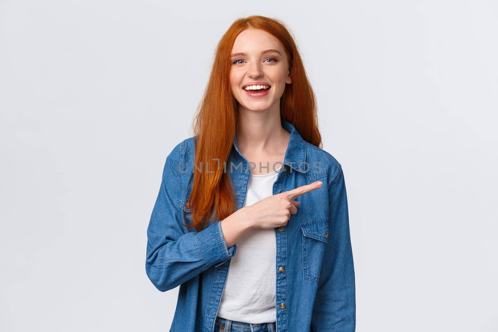 Carefree, friendly and happy outgoing redhead woman in denim shirt, pointing finger right and laughing at camera with beaming smile, standing white background, having discussion, communicate team.