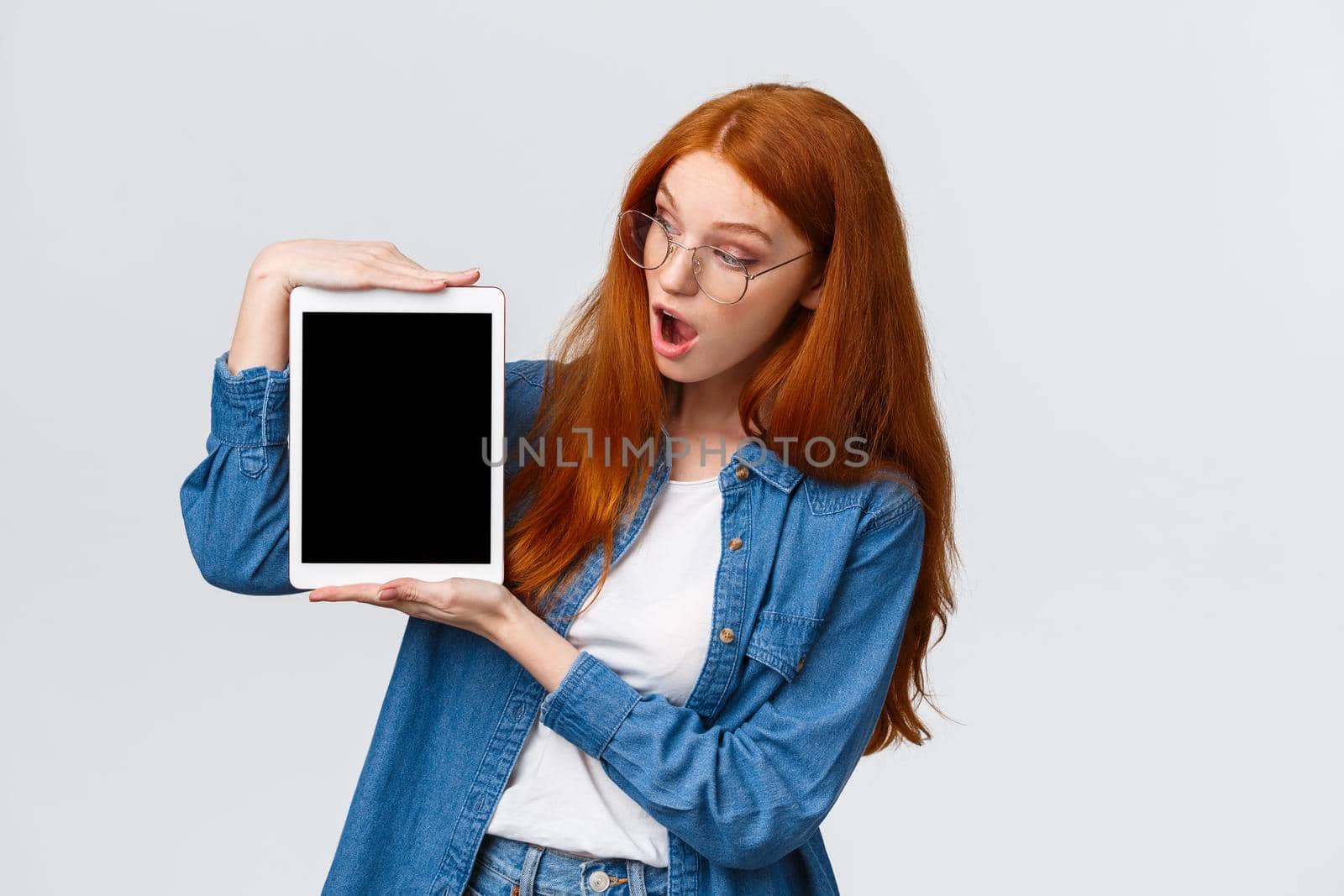 Surprised and curious cute redhead female in denim shirt, looking astonished digital tablet screen, say wow, checking out something awesome, standing white background. Copy space