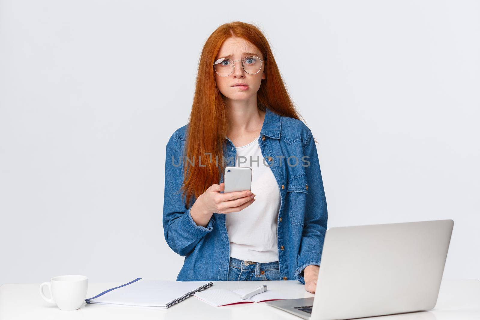 Anxious, worried cute redhead girl nervously looking camera and bite lip, have problem, cant meet deadline, holding smartphone, standing near laptop and working desk, white background.