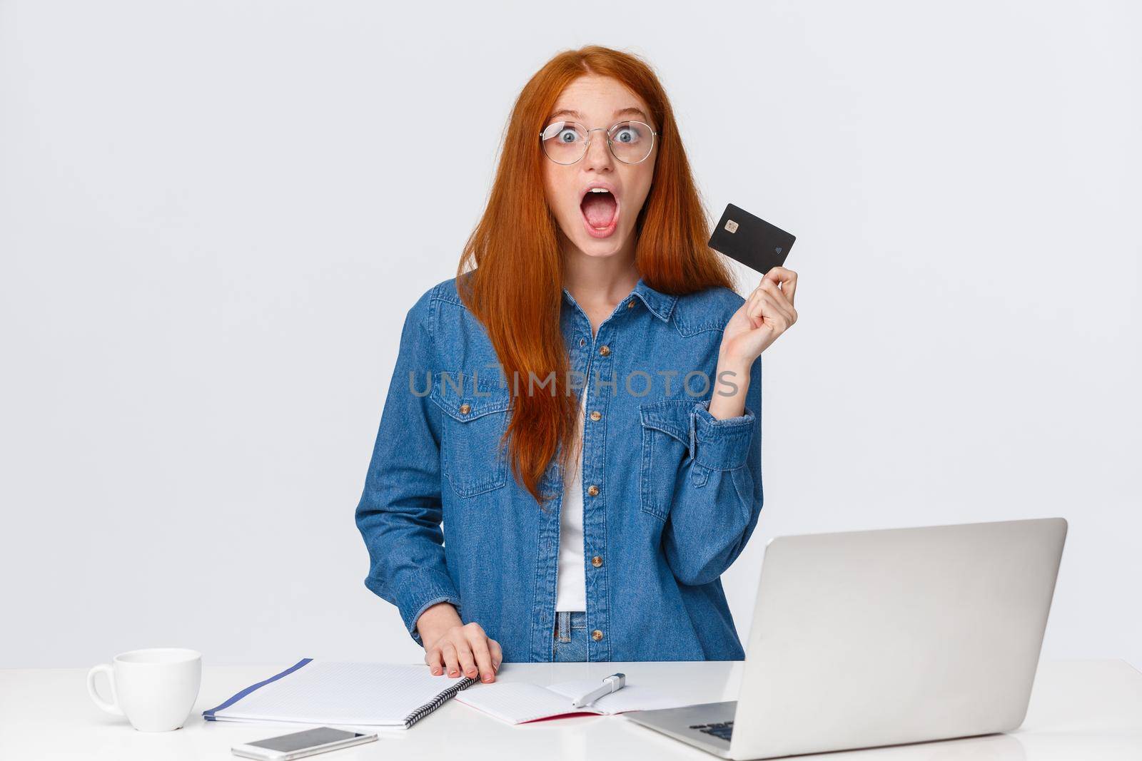 Excited and shook, impressed redhead girl holding credit card, got cashback for purchasing new furniture online store, visit website good prices, stand white background with laptop.