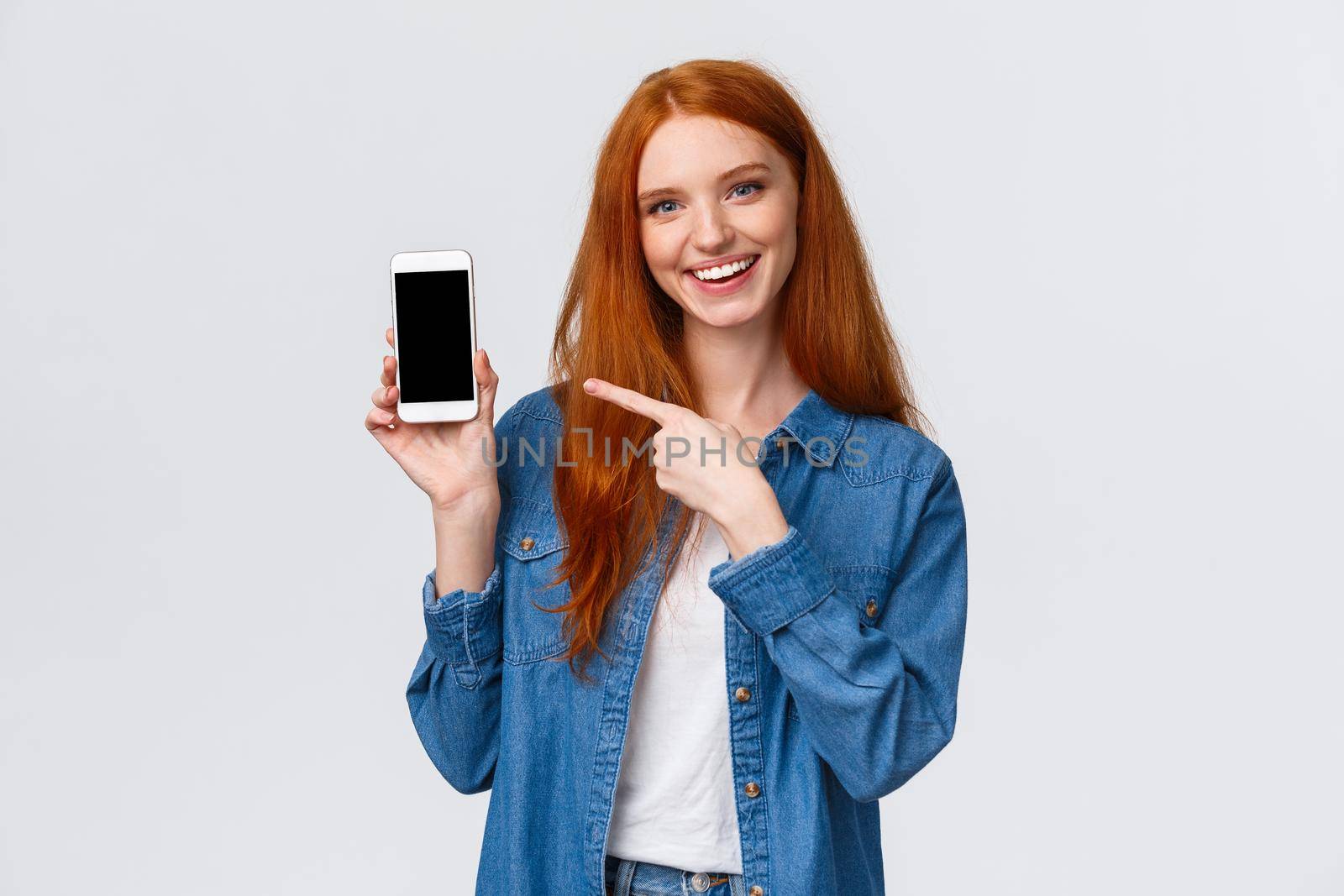 Waist-up portrait joyful charismatic redhead female review smartphone application, showing her photo with cute filter, pointing mobile display and smiling camera, white background.