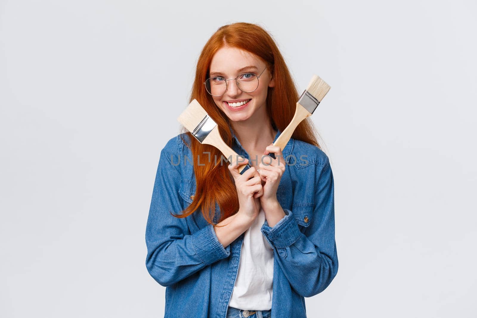 Dreamy and creative, joyful good-looking female designer, artist in glasses with red hair, holding two paintbrushes and smiling, plan repairment works, moving new house, white background.
