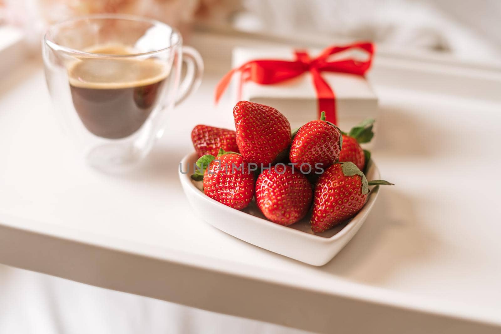 Breakfast for Valentine's Day. Heart shaped white plate with fresh strawberries, cup of coffee and flowers with gift in bed. Still life composition.