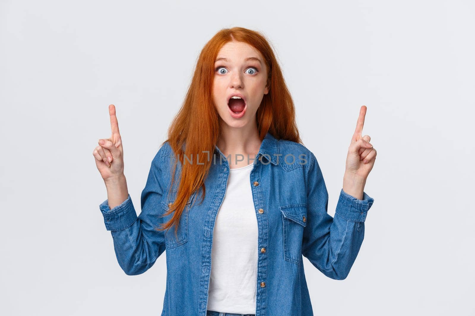 Astounded, fascinated good-looking caucasian woman with red hair, drop jaw, gasping saying wow staring camera with popped eyes, seeing something good and unbeliavable, pointing fingers up.