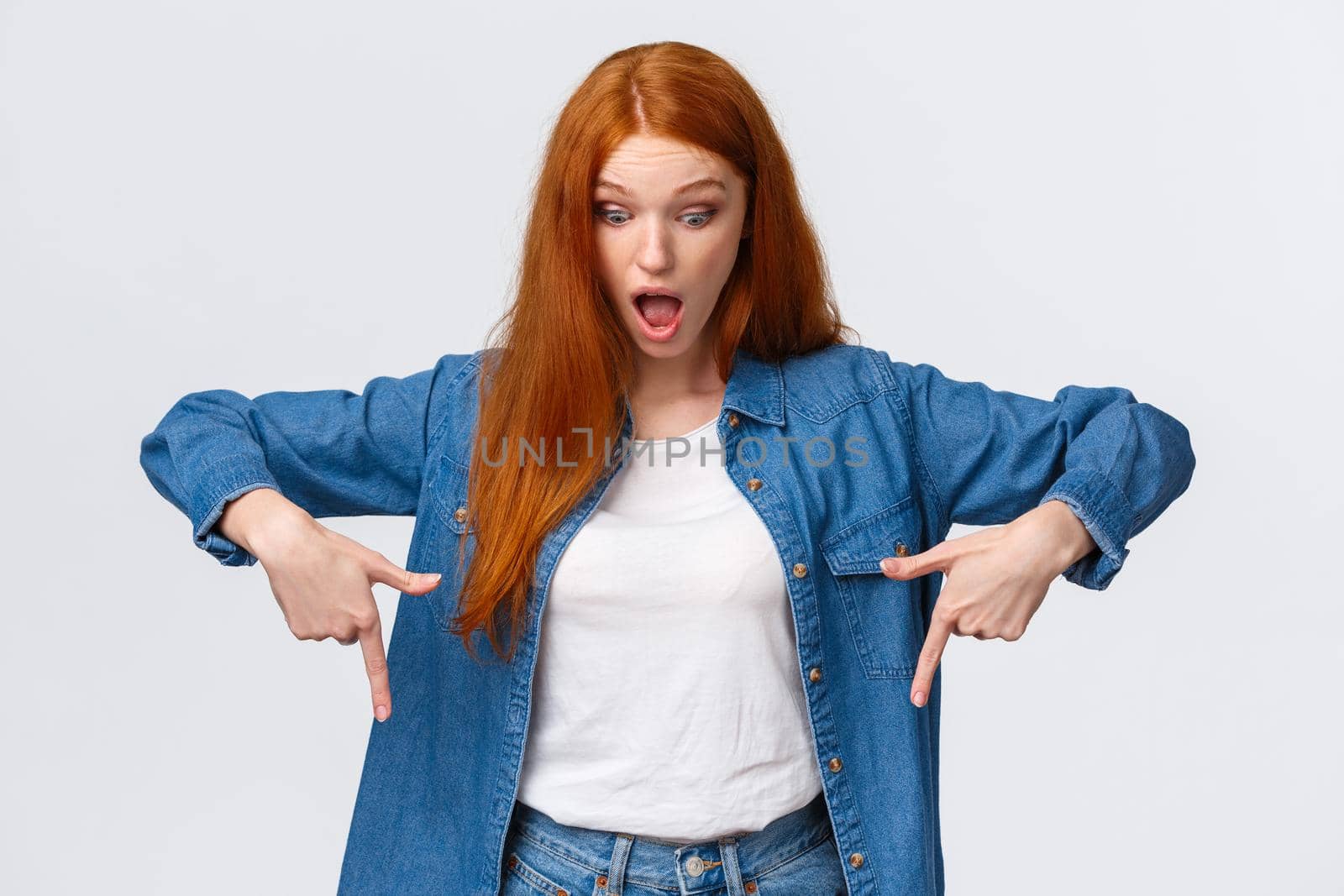 Waist-up portrait impressed, curious and astonished redhead cheerful girl in denim shirt, drop jaw, looking and pointing down starled speechless, standing white background amused by Benzoix