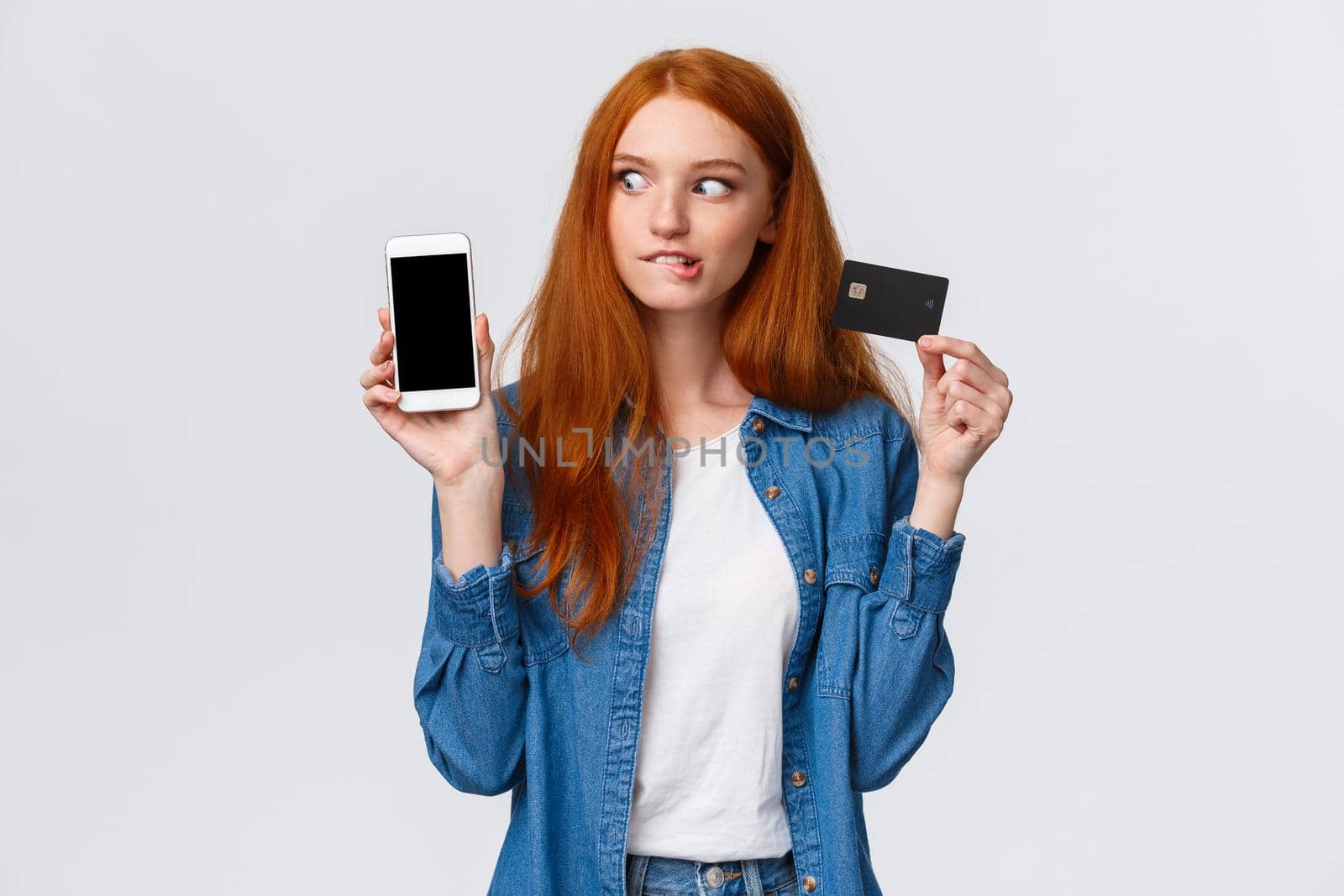 Girl consider buy thing with super good discount. Temped and wishful excited redhead female shopaholic looking at smartphone, shopping mobile app, holding credit card, bite lip eager purchase.