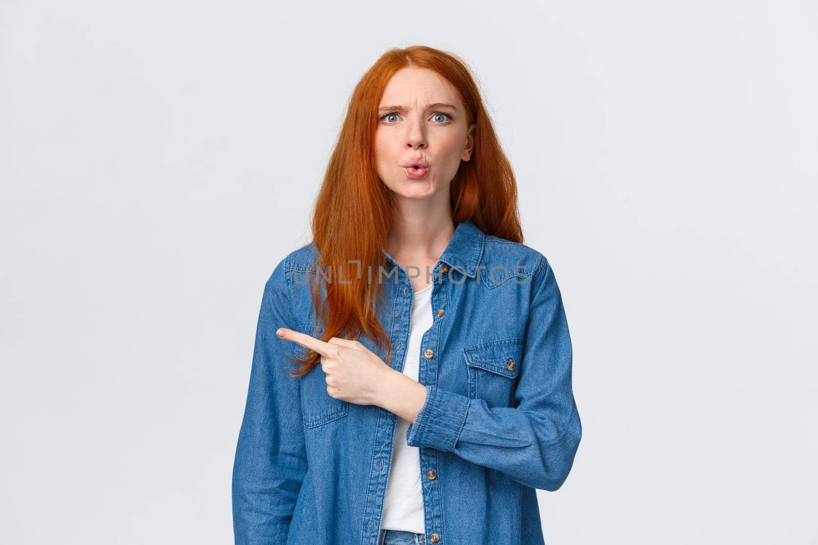 What is that. Alluring redhead girl in denim shirt, staring questioned, asking question and pointing finger left, frowning perplexed, dont know something, curious about product, white background.