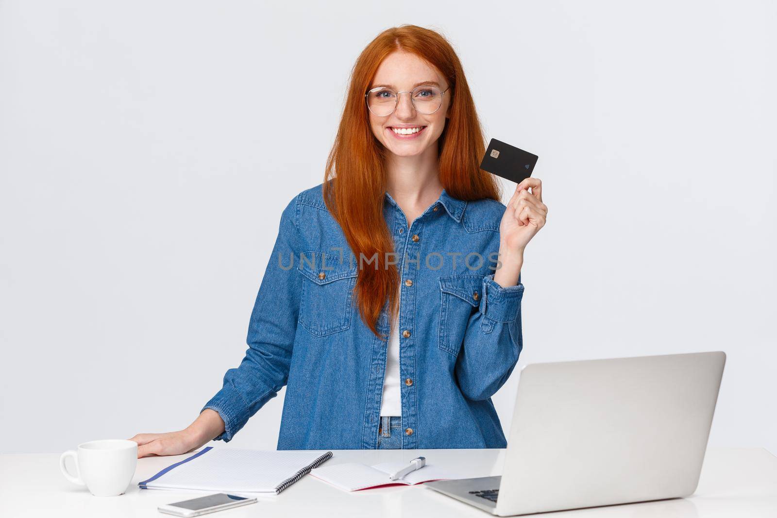 Cheerful good-looking girl dont need go shopping as purchase everything online, standing near table and laptop, showing credit card and smiling, recommend bank service, white background.