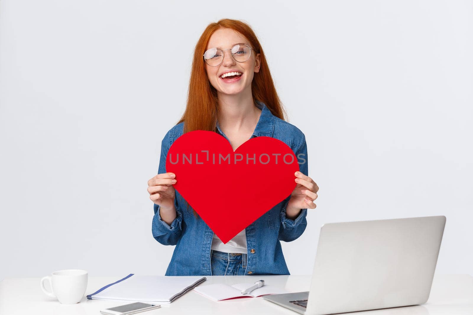 Valentines day, creativity and feelings concept. Cheerful smiling redhead girl with long-distance relatioship sending her love through internet, using web cam to show big red heart and say love you by Benzoix