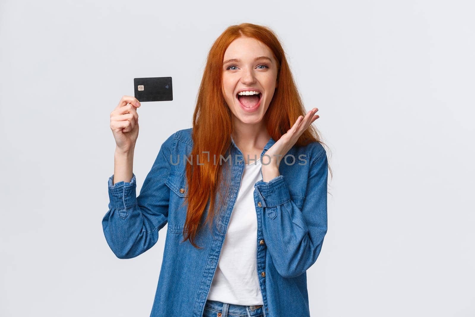 Charismatic happy good-looking redhead female student in casual clothes, holding credit card with beaming smile on face, astonished and pleased, receive salary, like bank offers, white background.