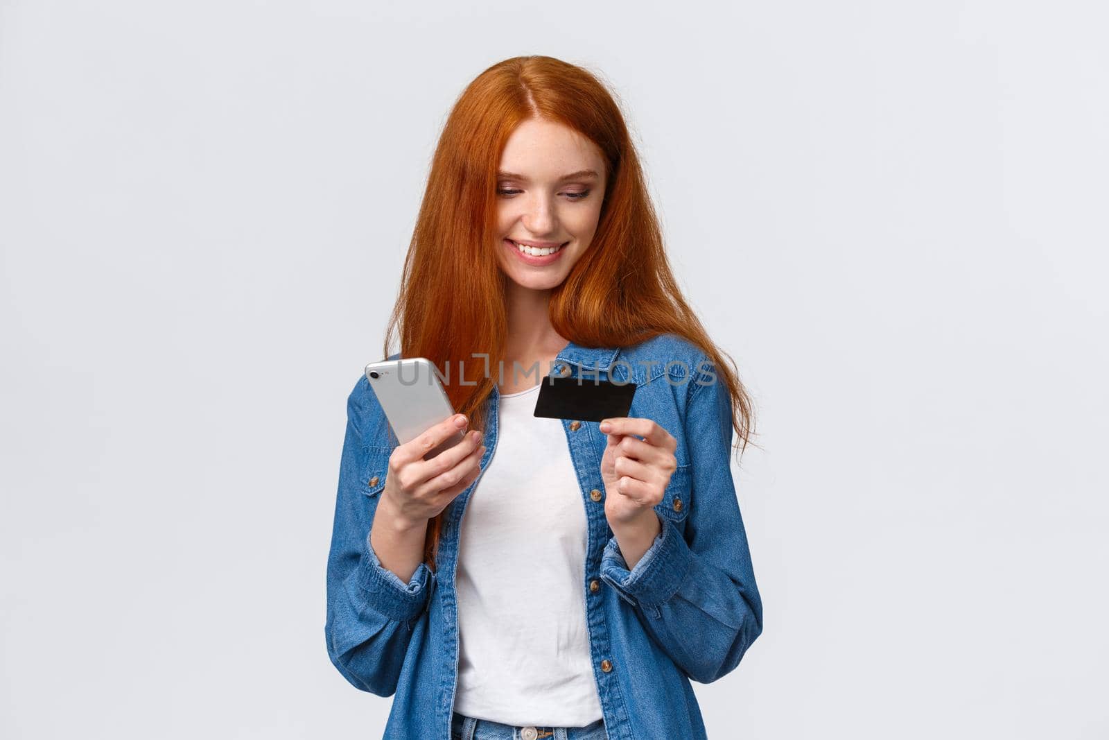Finance, business and education concept. Cheerful cute alluring redhead woman making online purchase, holding smartphone and credit card, smiling as buying present for valentines day internet store by Benzoix