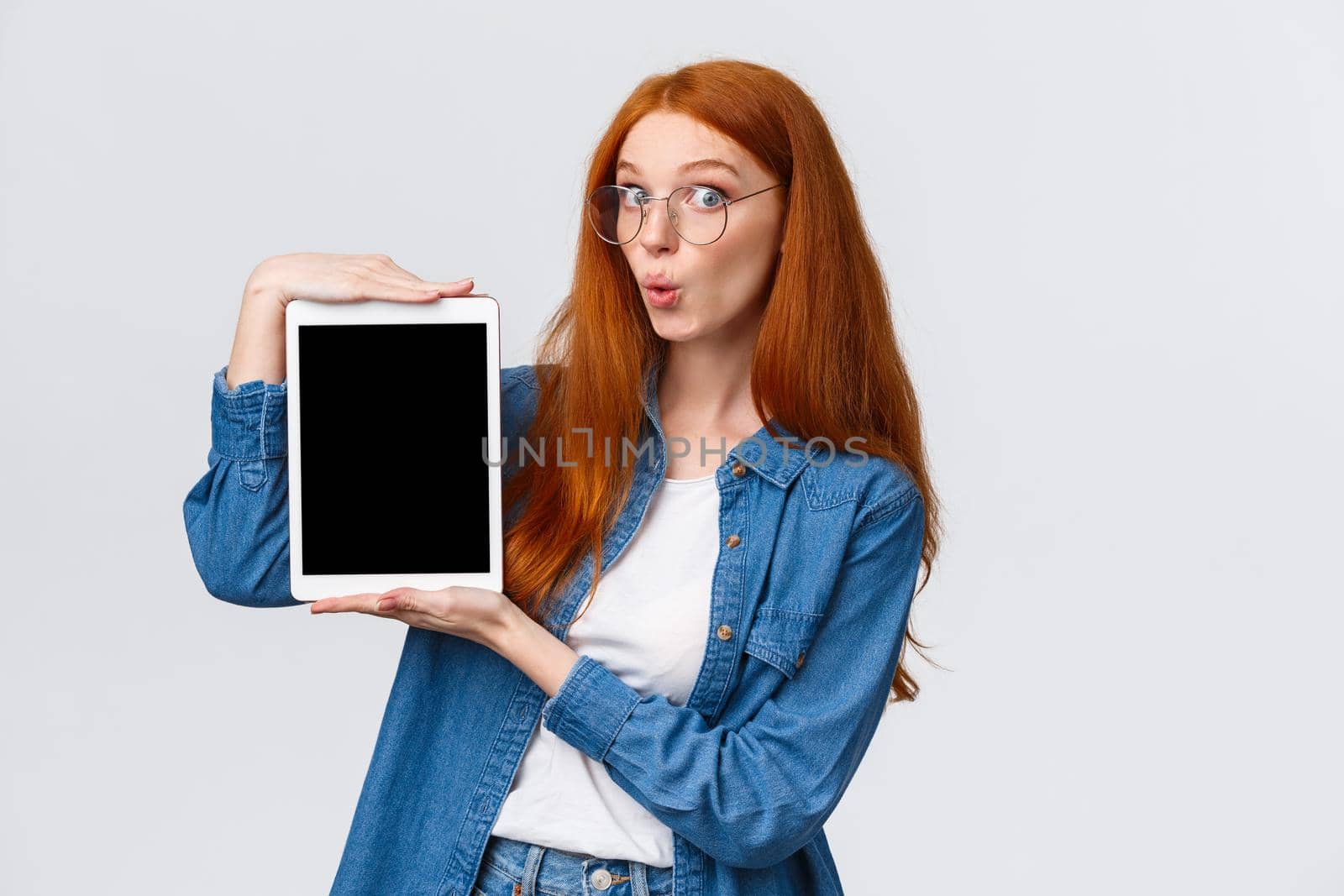 Amused, excited good-looking redhead woman in denim shirt, folding lips in wow sound, looking amazed camera, holding digital tablet, showing device screen, standing white background.