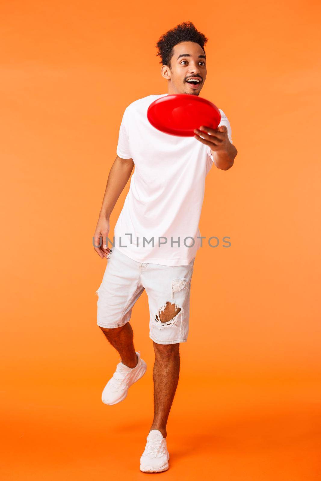 Full-length vertical shot carefree handsome african-american millennial guy enjoying summer leisure time, outdoor activity, throwing frisbee smiling and looking at friend, orange background.